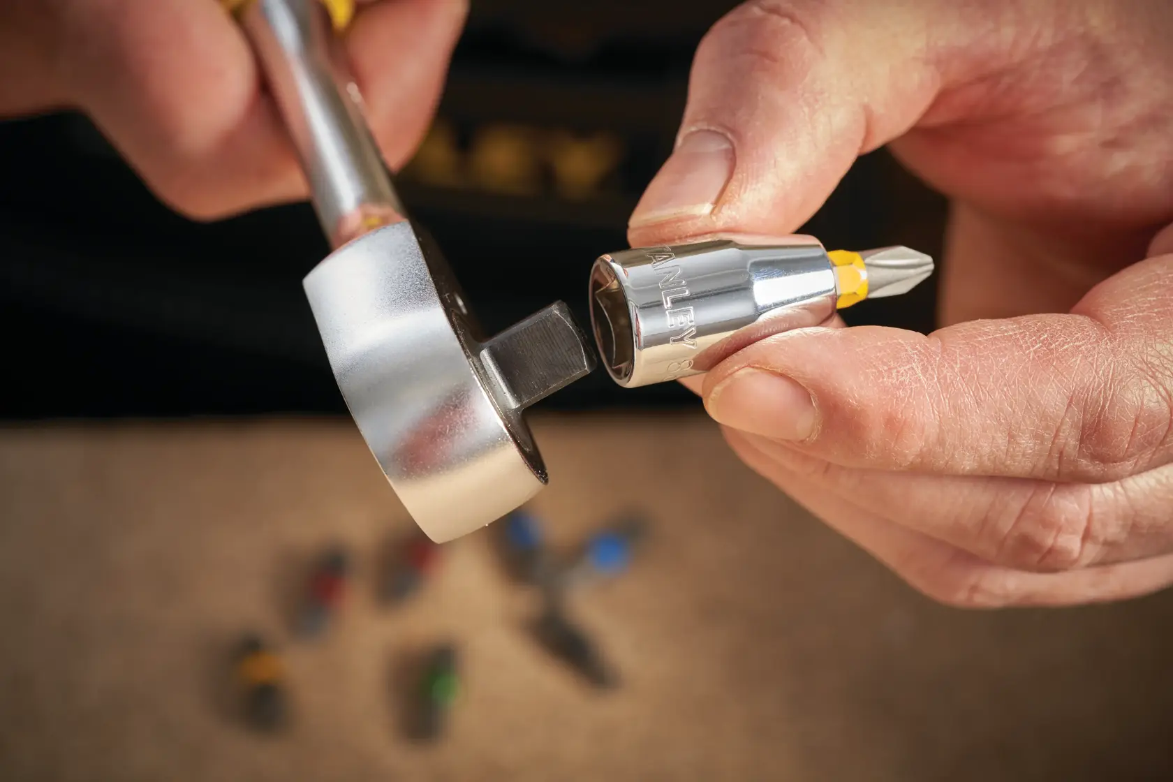 STANLEY STMT86250-0 carraca con adaptador de punta para destornillador en mano, primer plano, juego de herramientas al fondo.