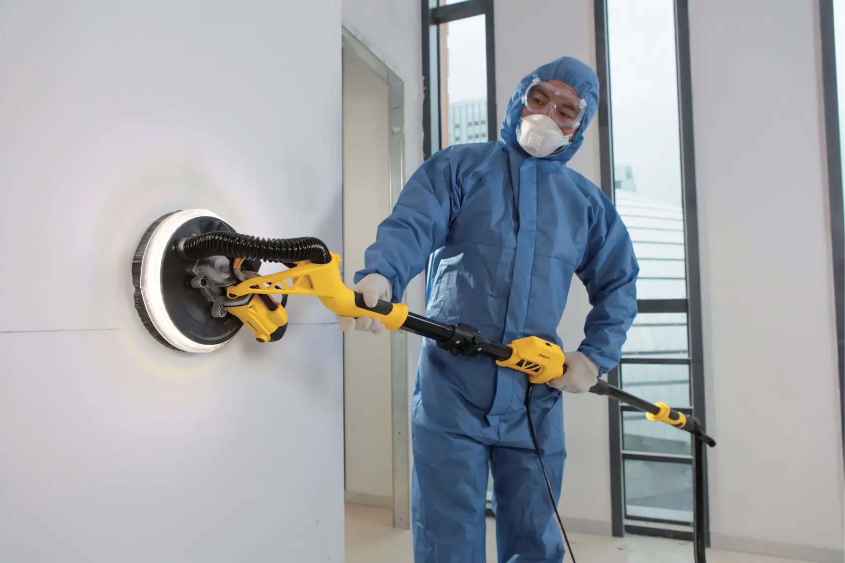 STANLEY SW75-BR lijadora de paneles de yeso en uso, herramienta amarilla lijando pared, persona con overol azul.