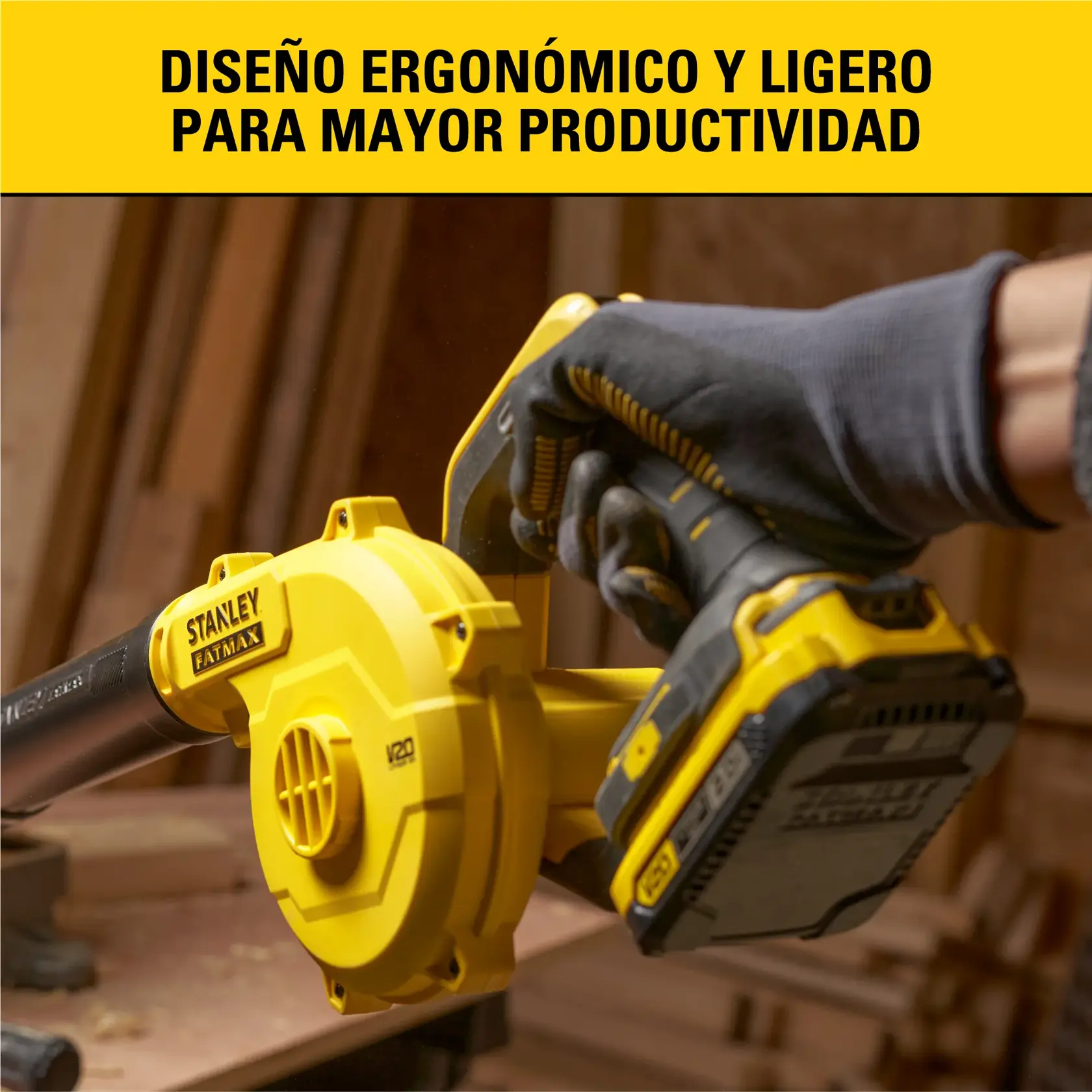 Vista en primer plano de una persona usando un soplador inalámbrico Stanley Fatmax V20 de color amarillo en un taller de carpintería. La persona lleva guantes de protección y sostiene la herramienta eléctrica ergonómica y ligera. Al fondo se ven tablas de madera y equipo de taller. La imagen incluye el texto en español 'Diseño ergonómico y ligero para mayor productividad' en una franja amarilla en la parte superior.