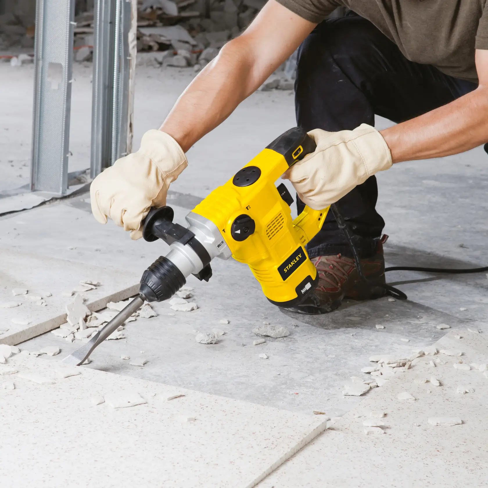 Una persona con guantes utiliza un martillo rotativo amarillo STANLEY (modelo STHR1232K) para romper baldosas en el suelo de una obra.