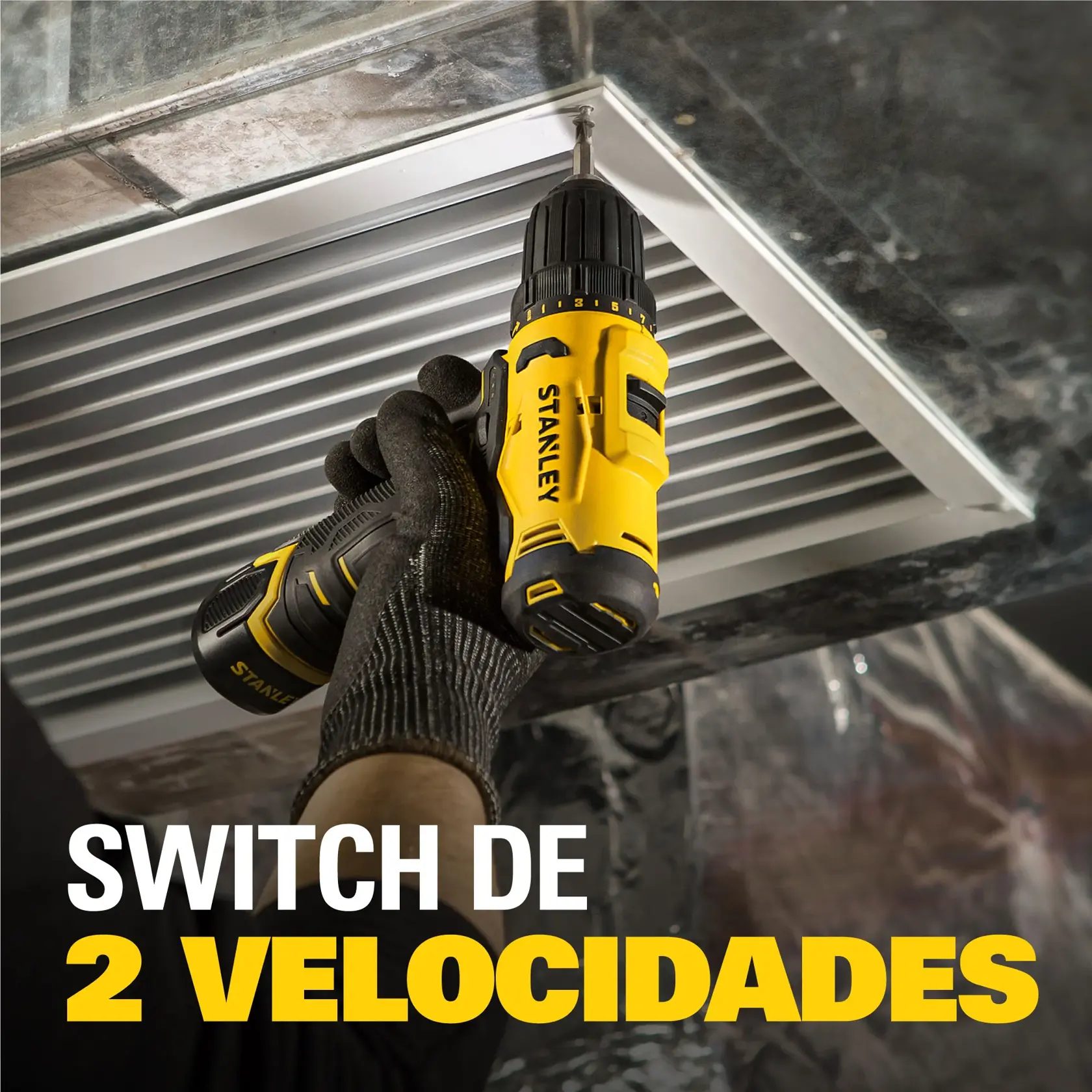Una persona con guante utiliza un taladro inalámbrico amarillo y negro STANLEY para atornillar en una rejilla metálica en el techo. El texto en la imagen dice: 'Switch de 2 velocidades'.