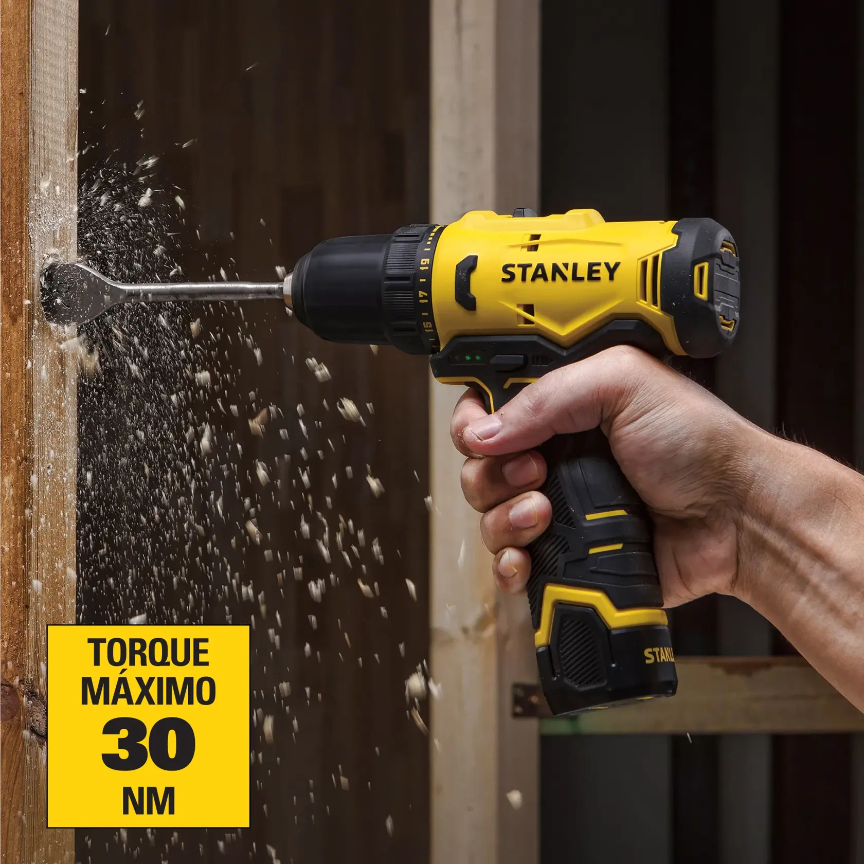 Una mano sostiene un taladro inalámbrico STANLEY amarillo y negro mientras perfora una viga de madera, con virutas de madera volando. La etiqueta del taladro resalta un torque máximo de 30 Nm.