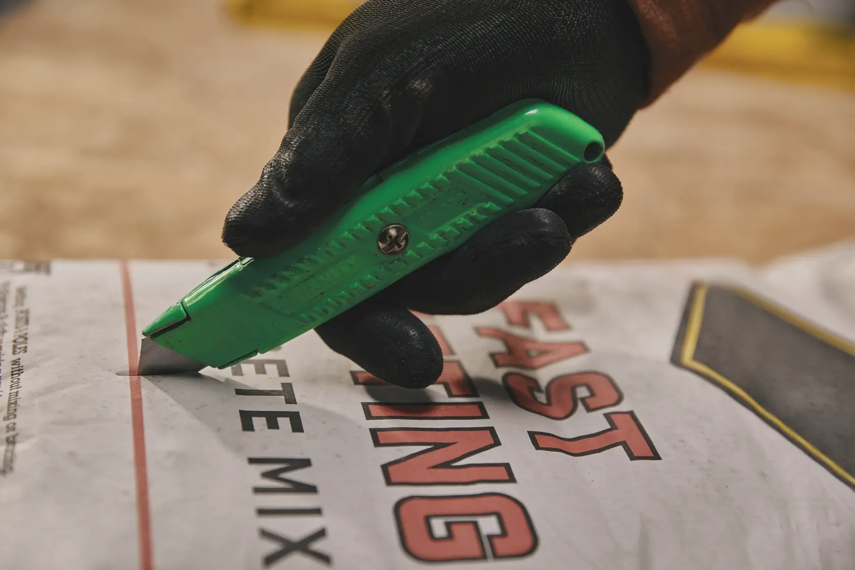 Cuchillo utilitario verde Stanley 10-179 cortando bolsa de mezcla de concreto, primer plano, sujetado con guante negro.