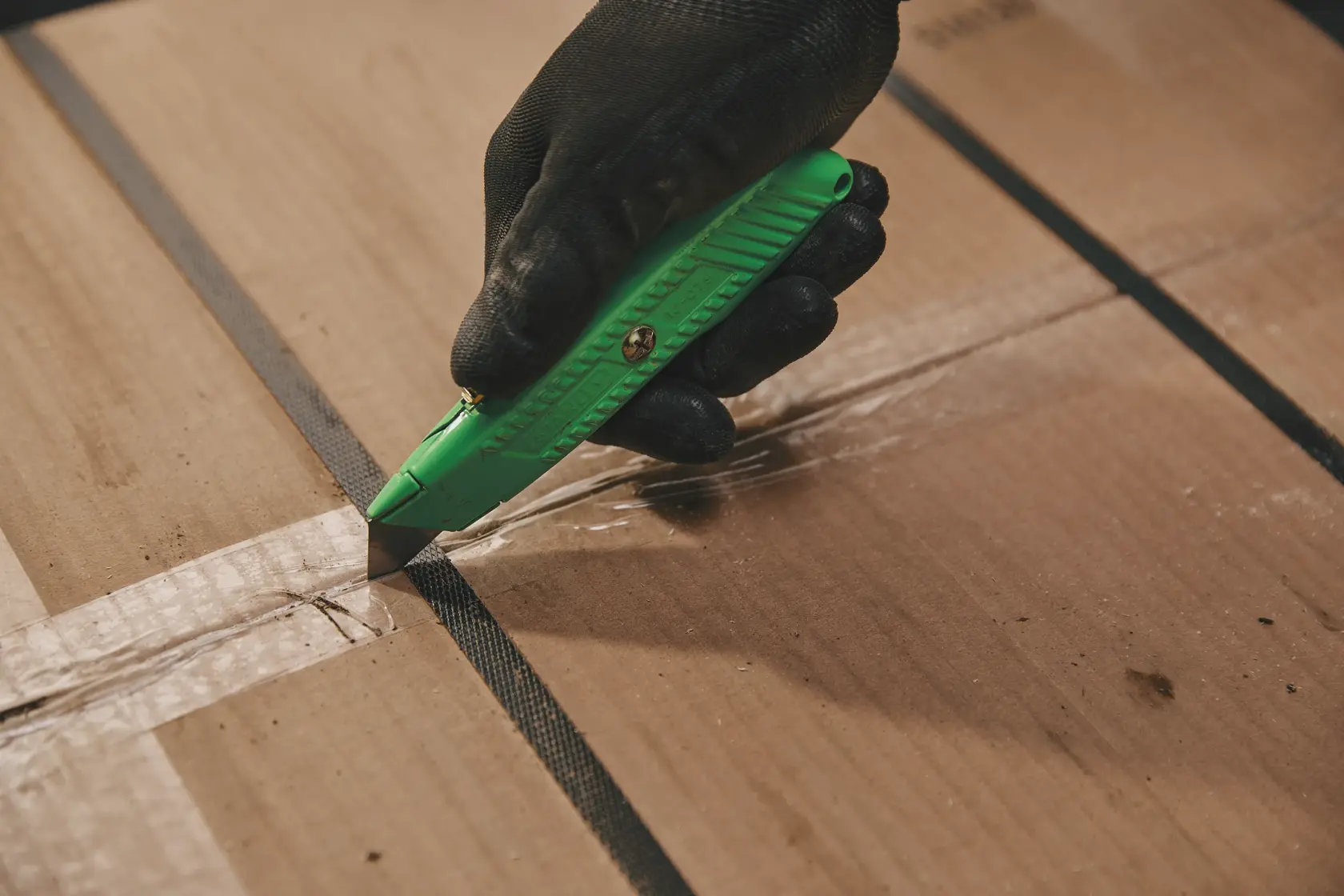 Foto del cúter STANLEY 10-179 cortando cinta en una caja de cartón, mano enguantada sujetando el cúter verde.