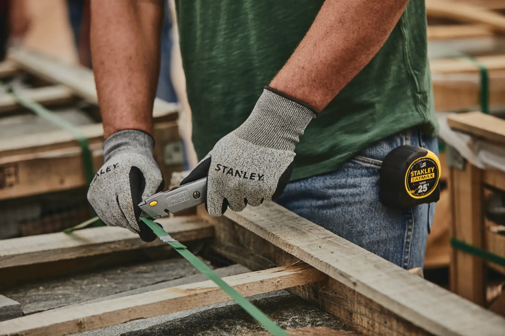 Stanley 10-499 cuchillo utilitario en uso, trabajador con guantes cortando fleje verde en palé de madera.