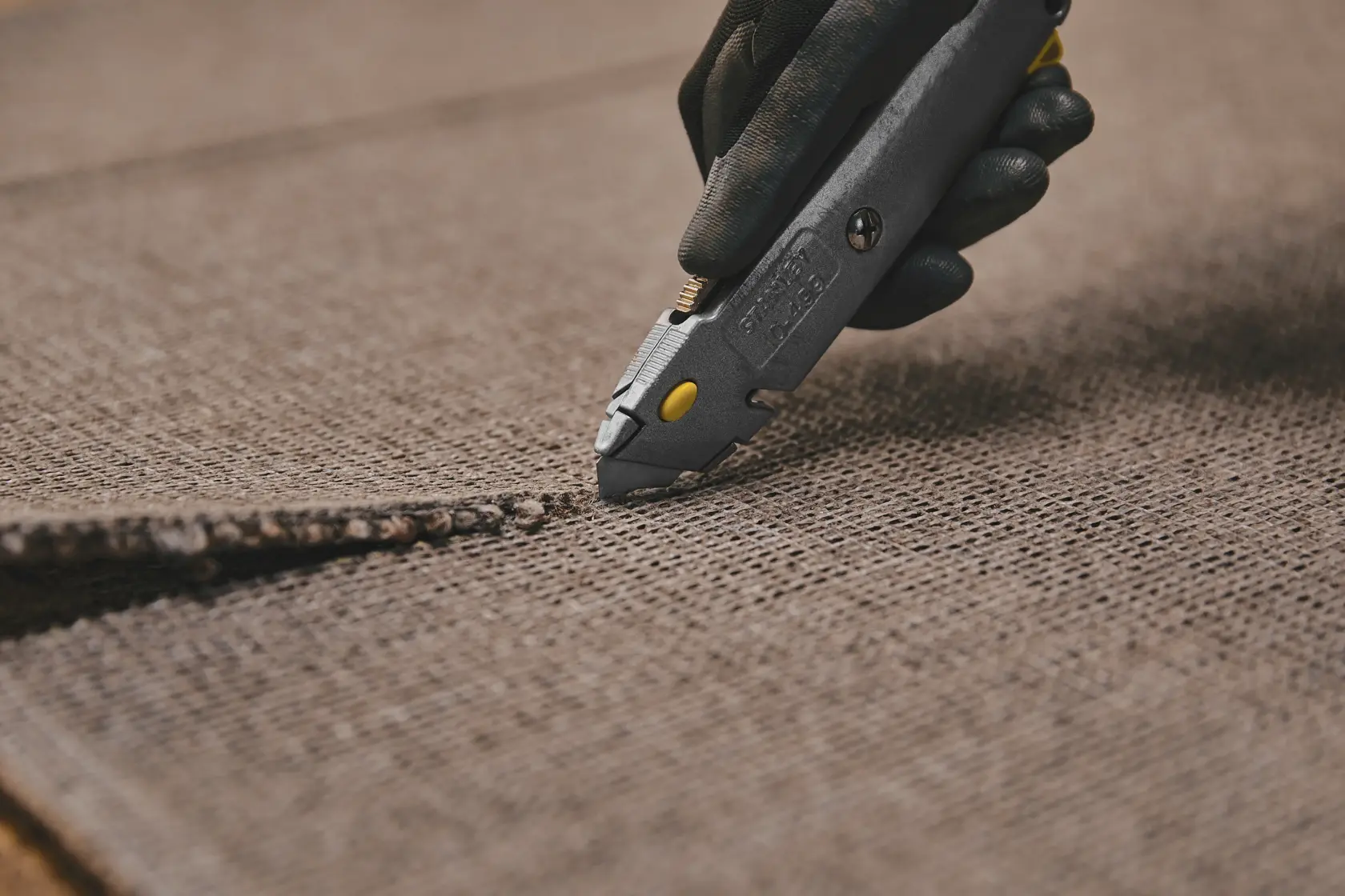 Fotografía del cuchillo multiuso STANLEY 10-499 cortando alfombra marrón, vista de cerca de la hoja en uso.