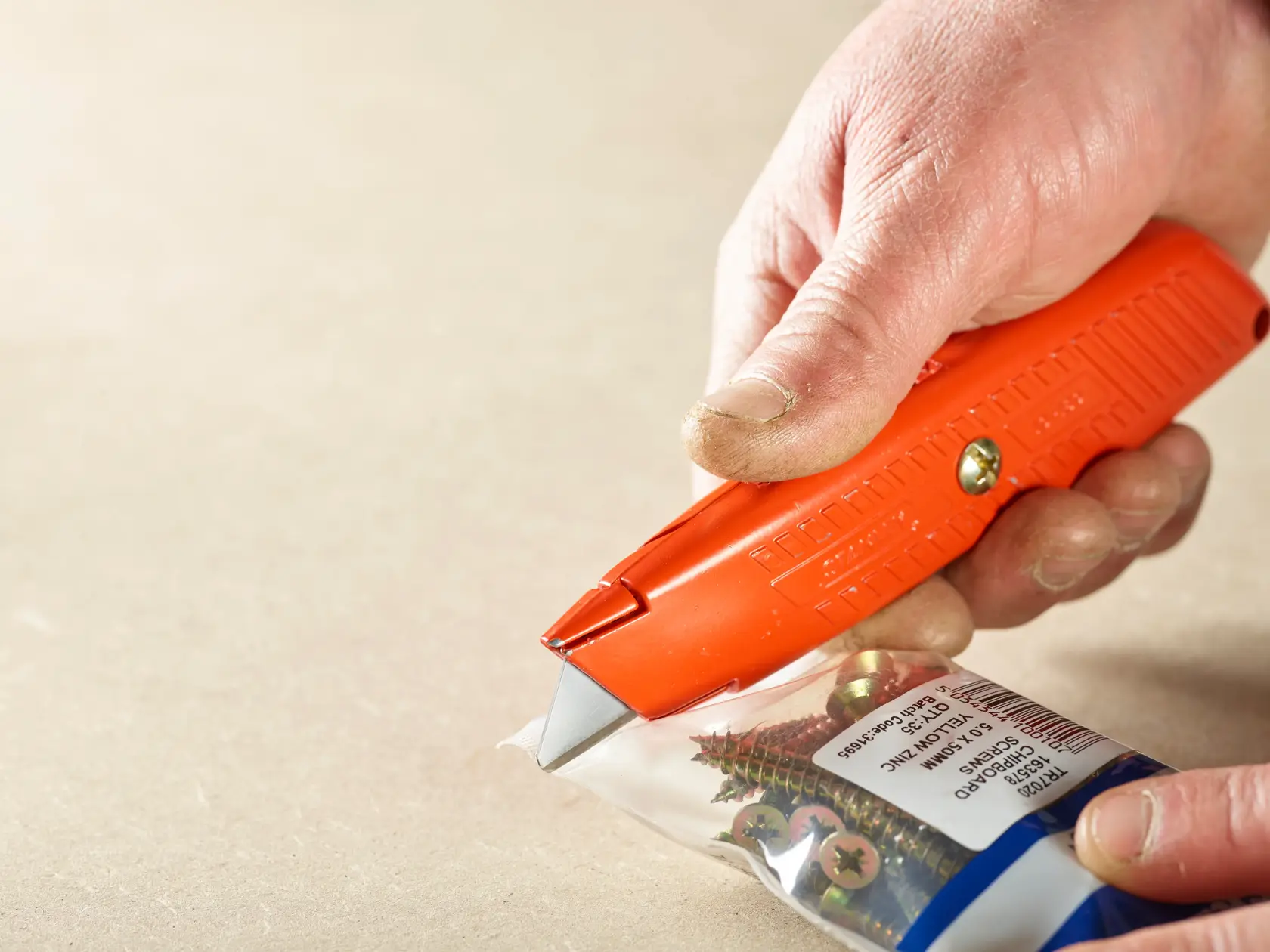 Cuchillo STANLEY 2-11-987 cortando paquete plástico de tornillos, primer plano del mango naranja y la hoja.