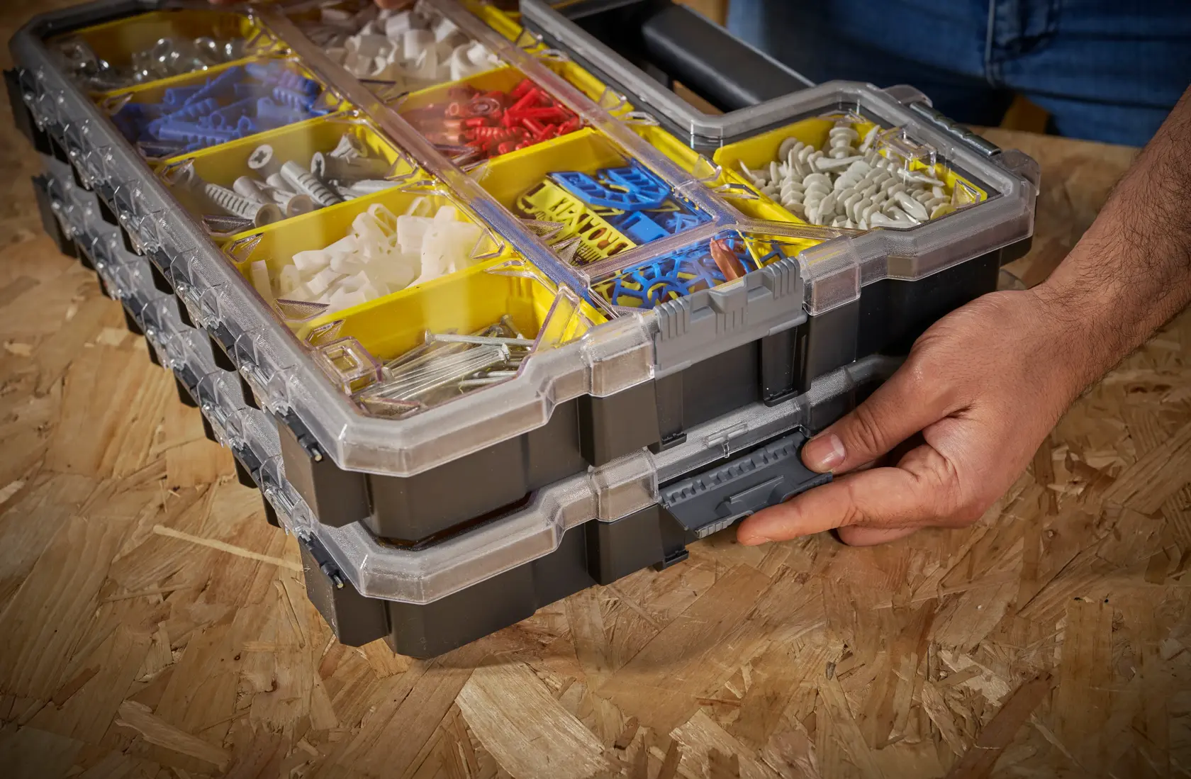 Caja organizadora STANLEY FMST14820 con compartimentos amarillos, tapa transparente y sujetadores surtidos, mano abriendo el clip frontal.