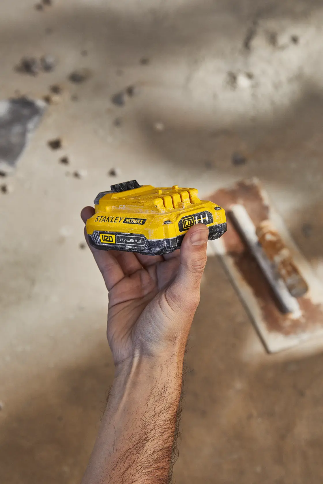 Foto de una batería STANLEY SB201-B2 V20 de litio color amarillo y negro sostenida en mano, fondo de obra.