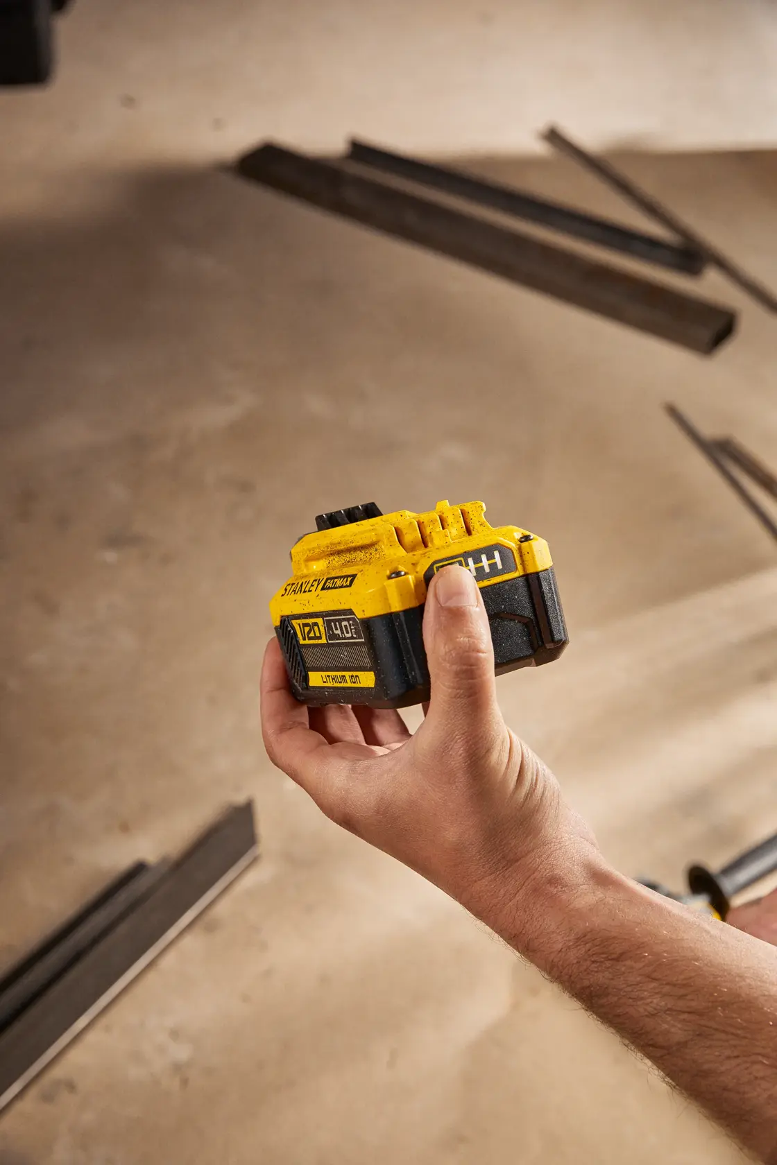 Fotografía de batería STANLEY SB204-KR 20V de ion de litio sostenida en una mano, materiales de construcción al fondo.