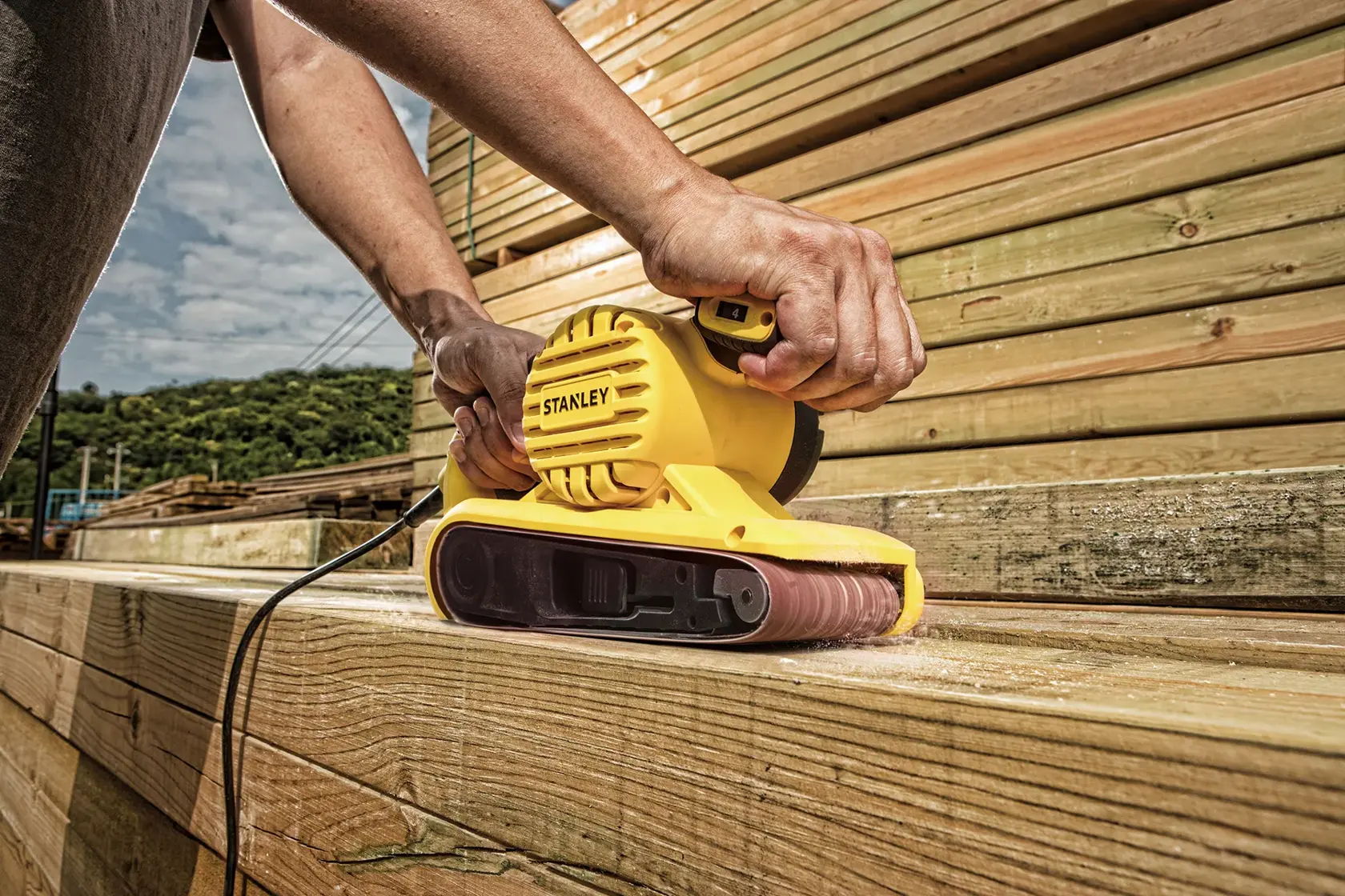 Lijadora de banda Stanley SB90-B2C amarilla utilizada en tablones de madera al aire libre, fotografía de cerca.