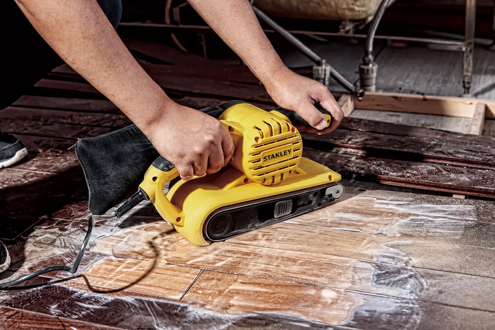 Lijadora de banda STANLEY SB90-B2C en uso sobre piso de madera, primer plano con bolsa de polvo y mango ergonómico.