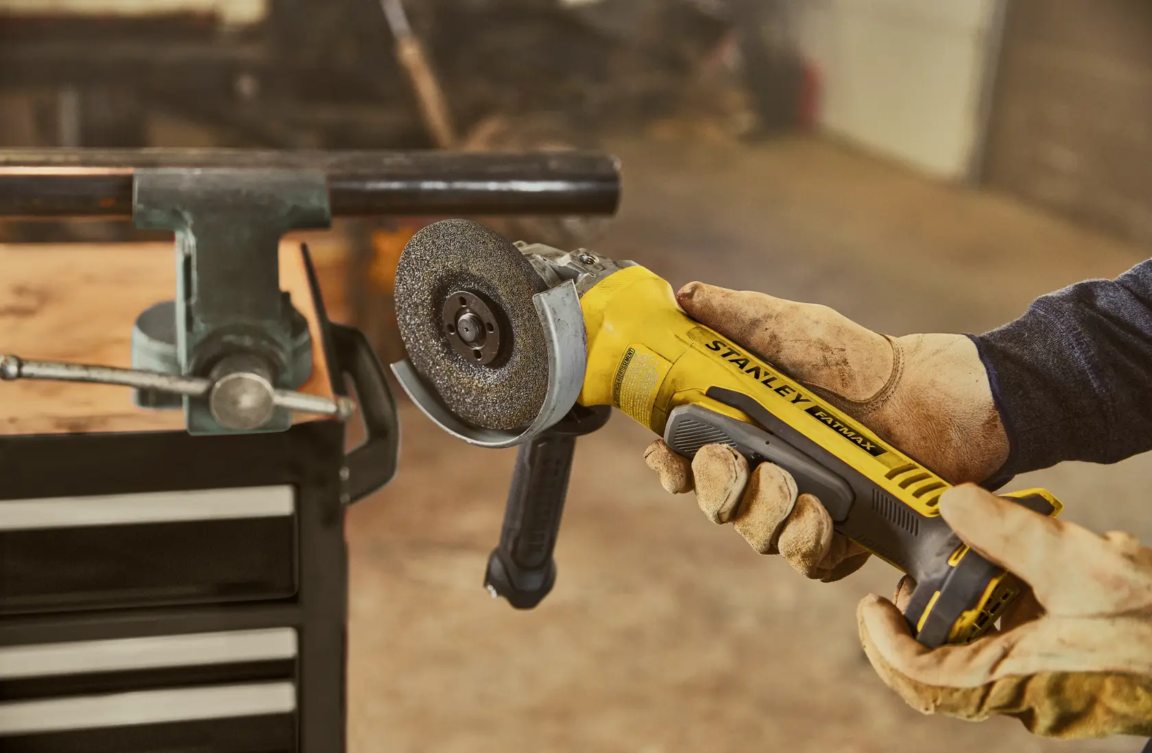 Stanley SBG700 amoladora angular sostenida por manos enguantadas cerca de tubo metálico en taller.