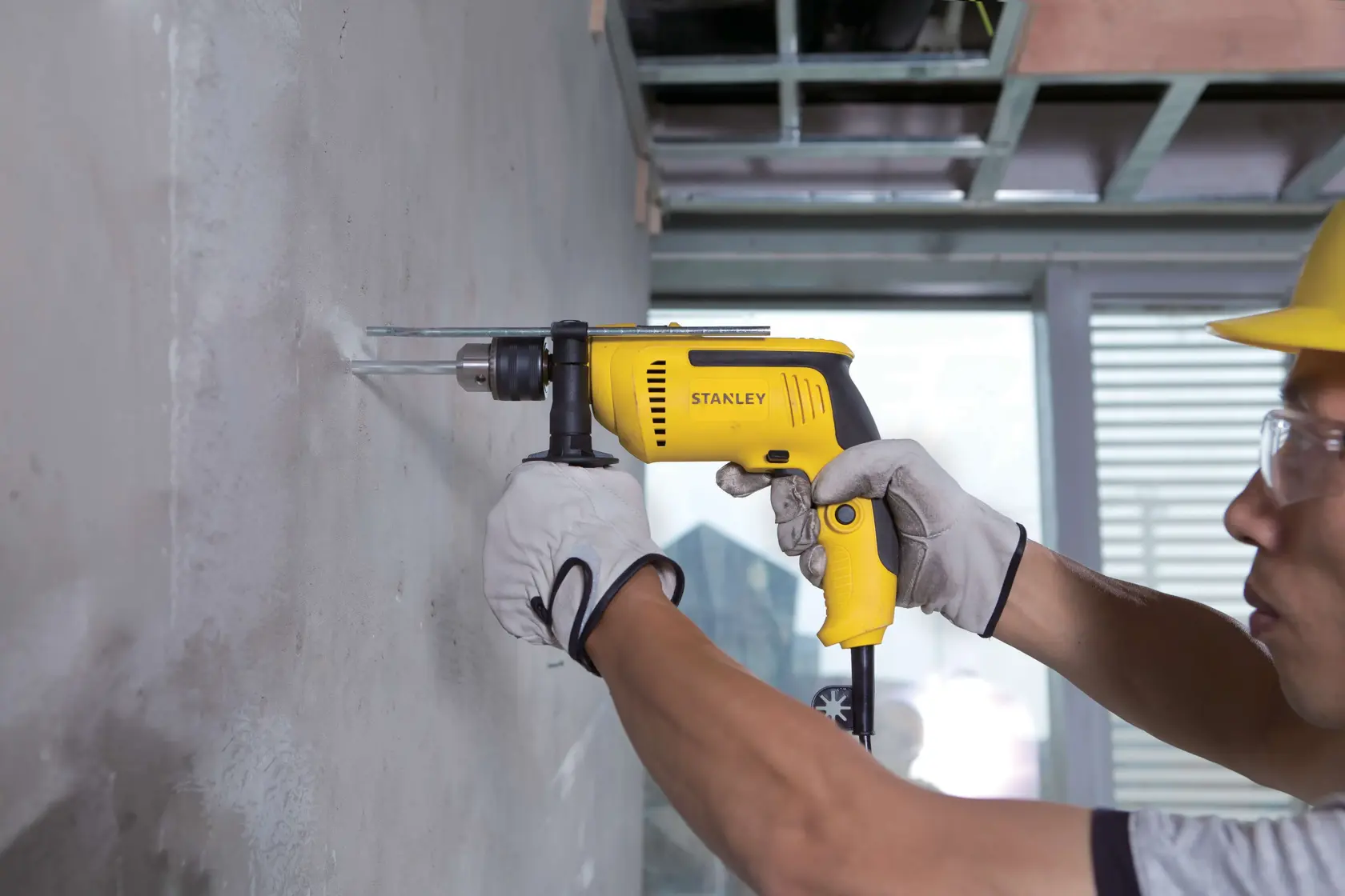 Fotografía de taladro amarillo STANLEY SDH700 con cable usado en pared de concreto por un trabajador con guantes.