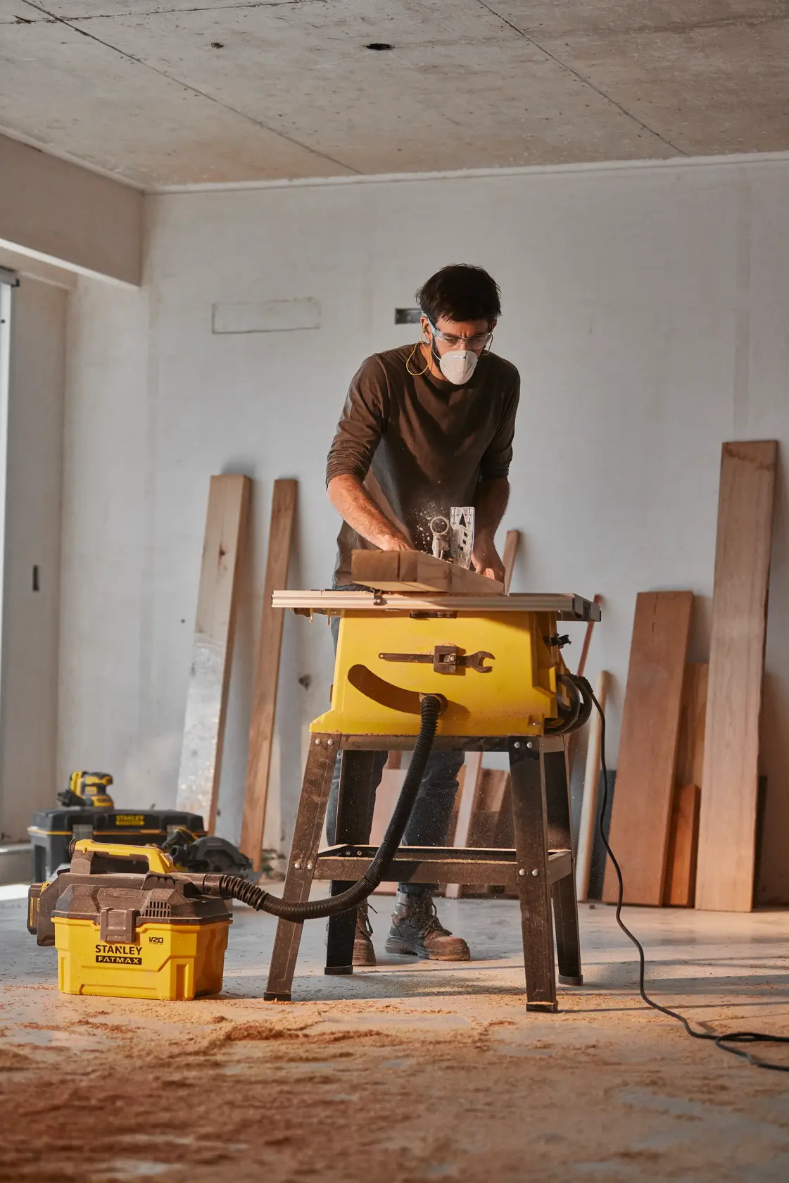 Aspiradora STANLEY SST2001-B2 conectada a una sierra de mesa en un taller con tablones de madera al fondo, vista lateral.