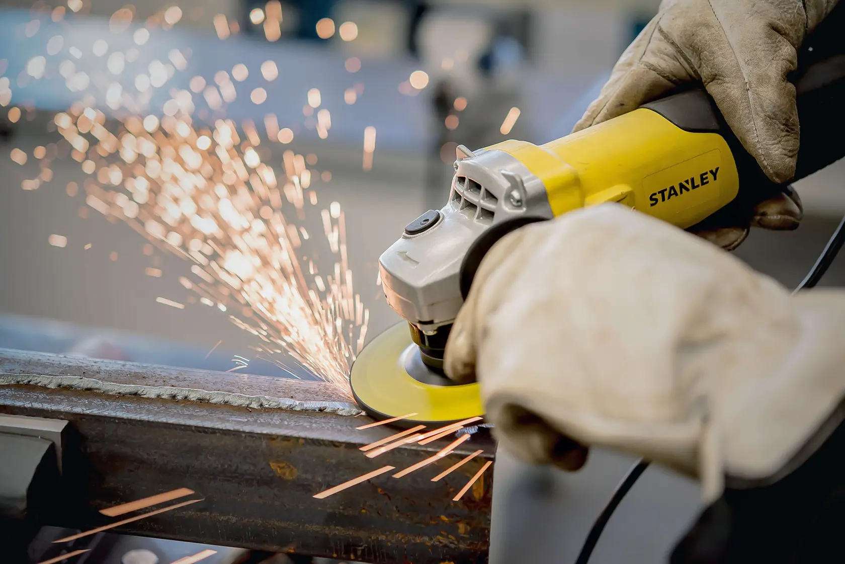 Fotografía de la amoladora angular STANLEY STA0413 en uso, lijando metal con chispas visibles, operada con guantes.