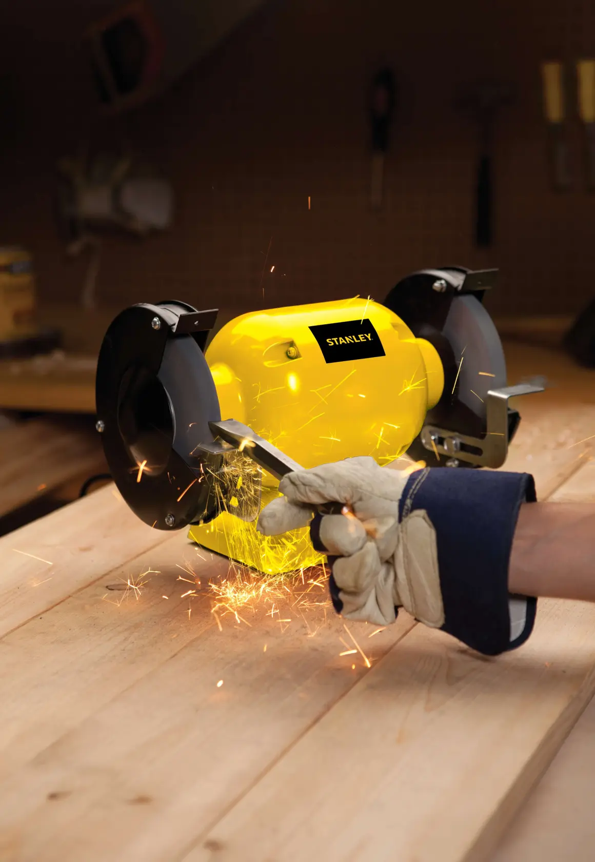 STANLEY STGB3715 esmeriladora de banco en uso sobre banco de trabajo de madera, chispas al afilar herramienta con guante.
