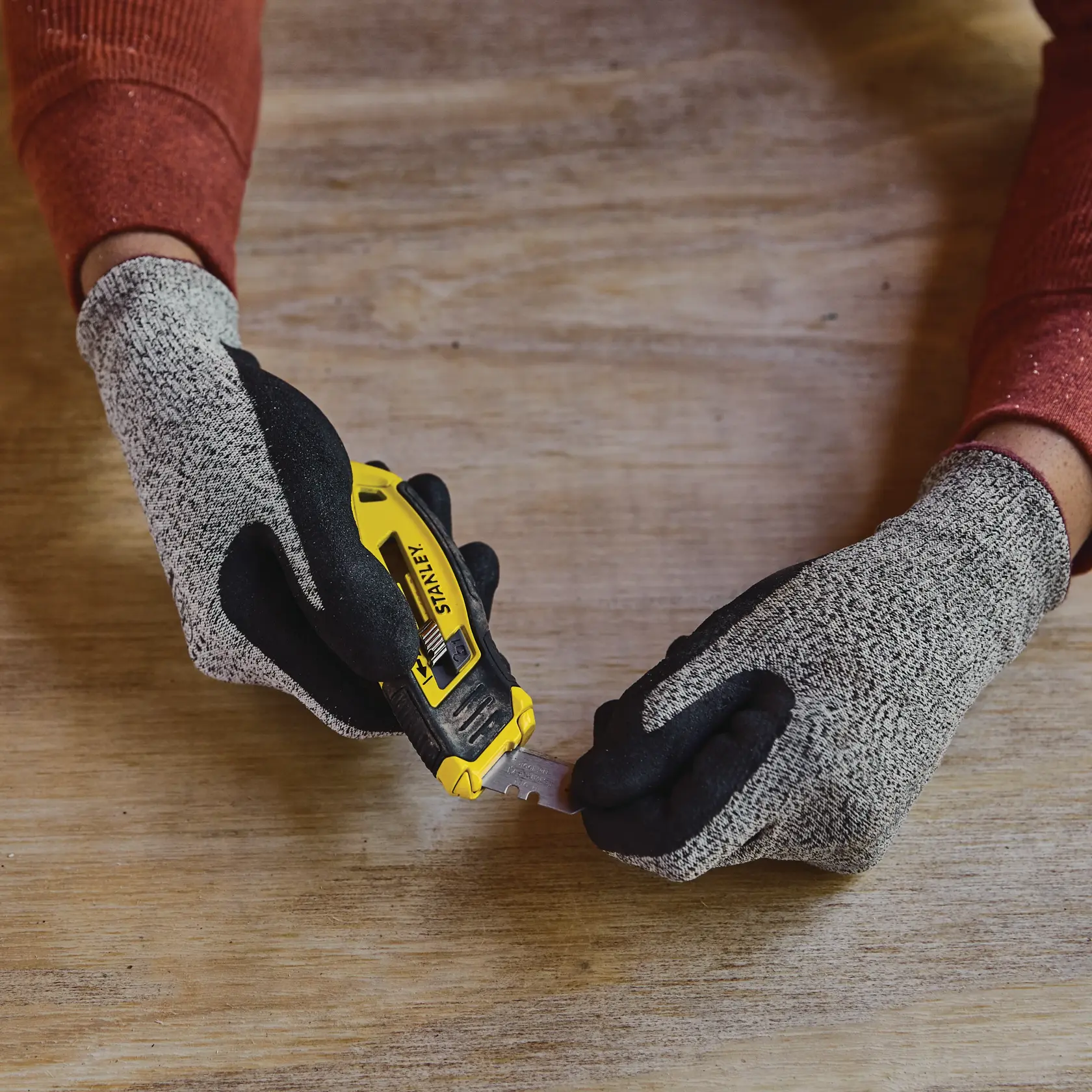 Fotografía de cuchillo utilitario STANLEY STHT10432 con mango amarillo y negro, sostenido por manos con guantes sobre superficie de madera.