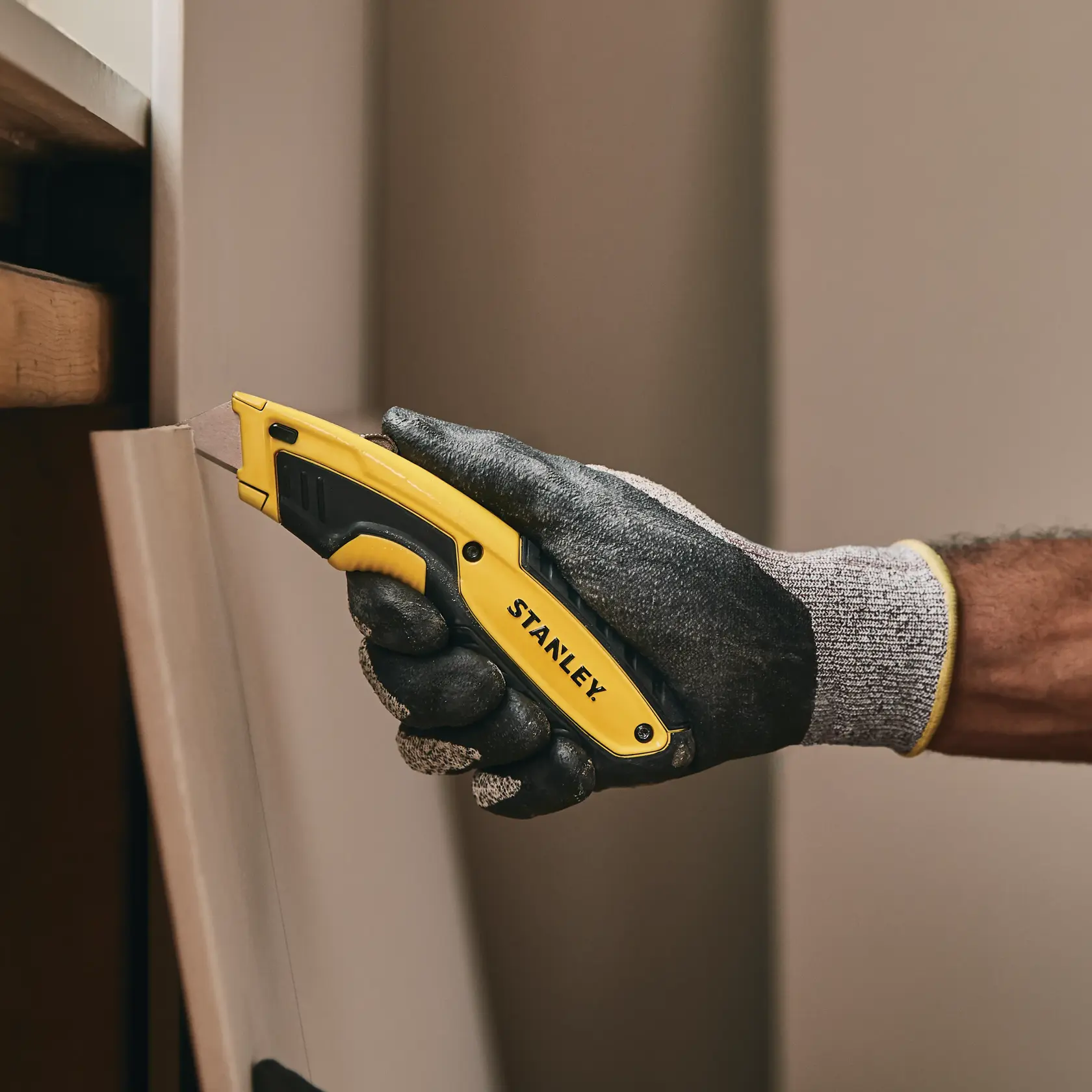 Cuchillo utilitario STANLEY STHT10479 con mango amarillo y negro, mano enguantada cortando panel de madera.