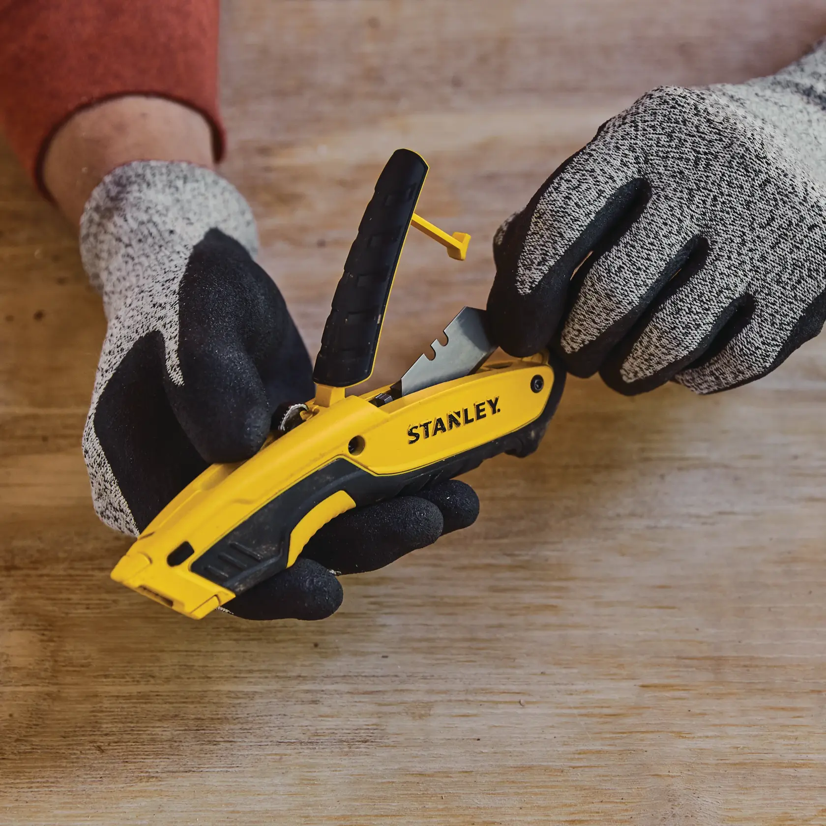 Fotografía del cuchillo utilitario amarillo y negro STANLEY STHT10479 con guantes, cambiando la hoja sobre superficie de madera.