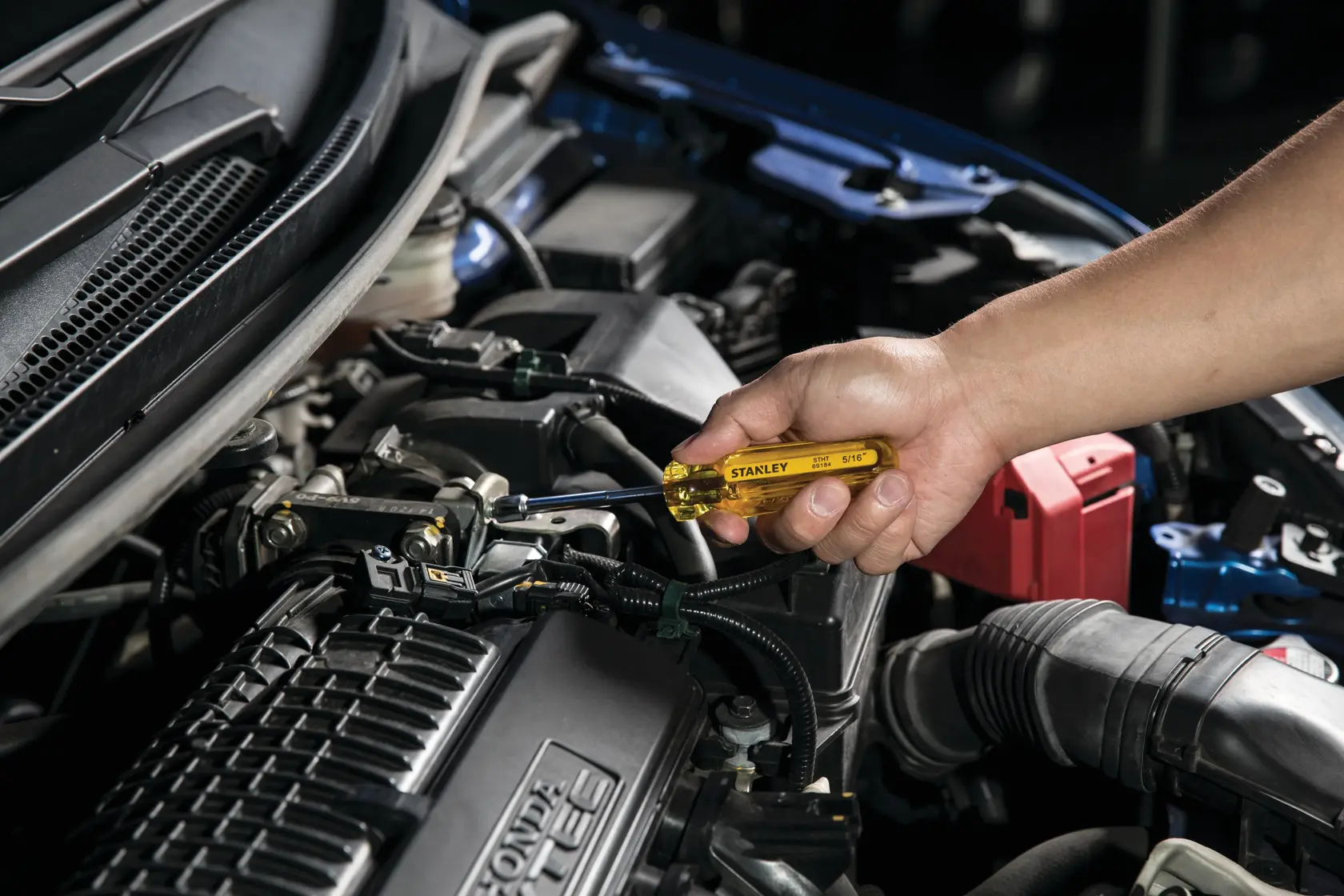Fotografía del destornillador STANLEY STHT69184 en uso sobre motor de automóvil, mano sosteniendo mango amarillo en primer plano.
