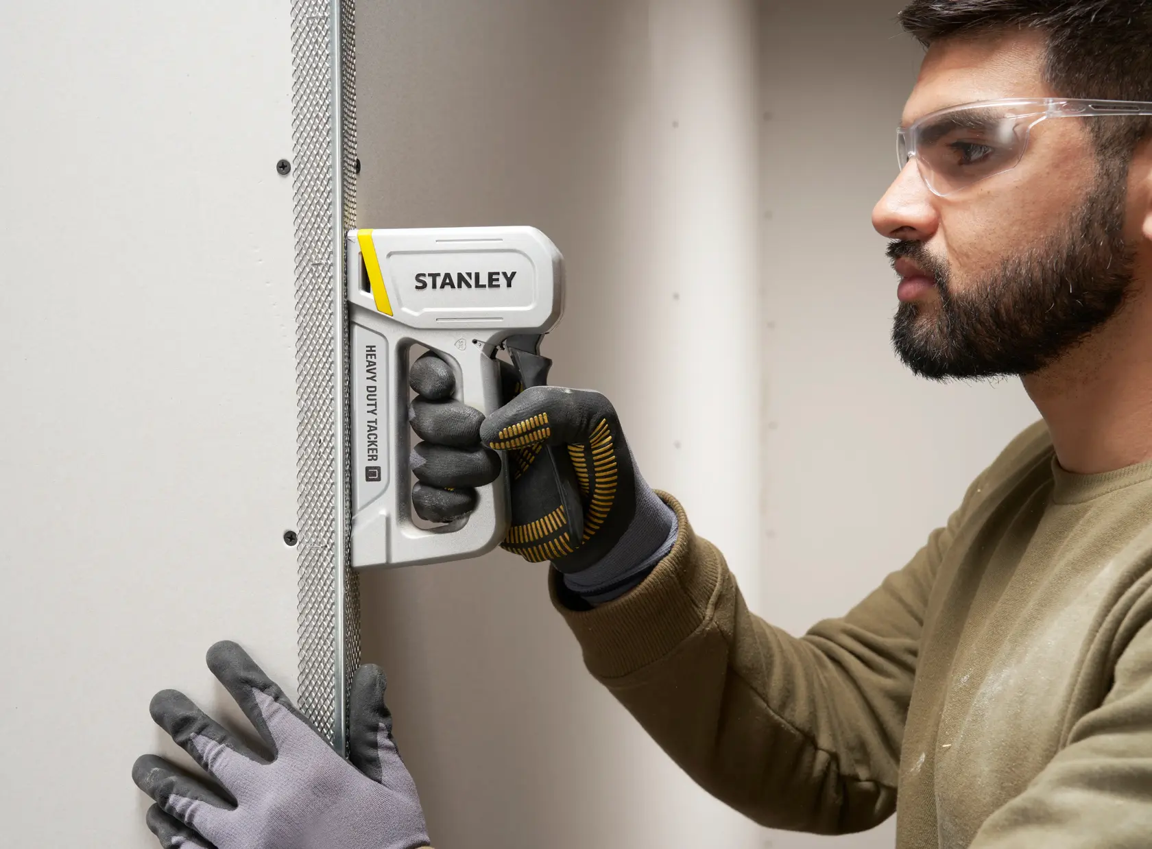 Fotografía de una persona usando una grapadora de gran resistencia STANLEY STHT70150 en panel de yeso, con guantes de trabajo.