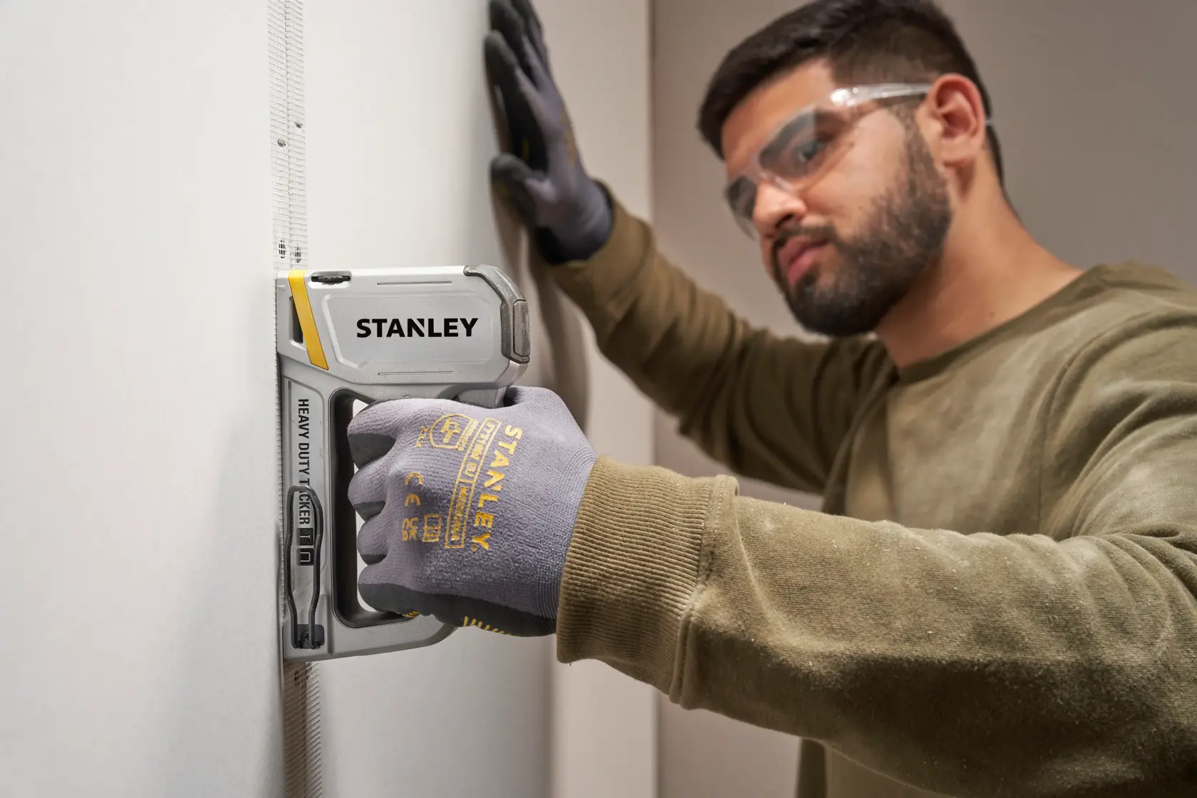 Fotografía de la grapadora Stanley STHT70250 de alta resistencia en uso, sostenida por mano enguantada en pared blanca.