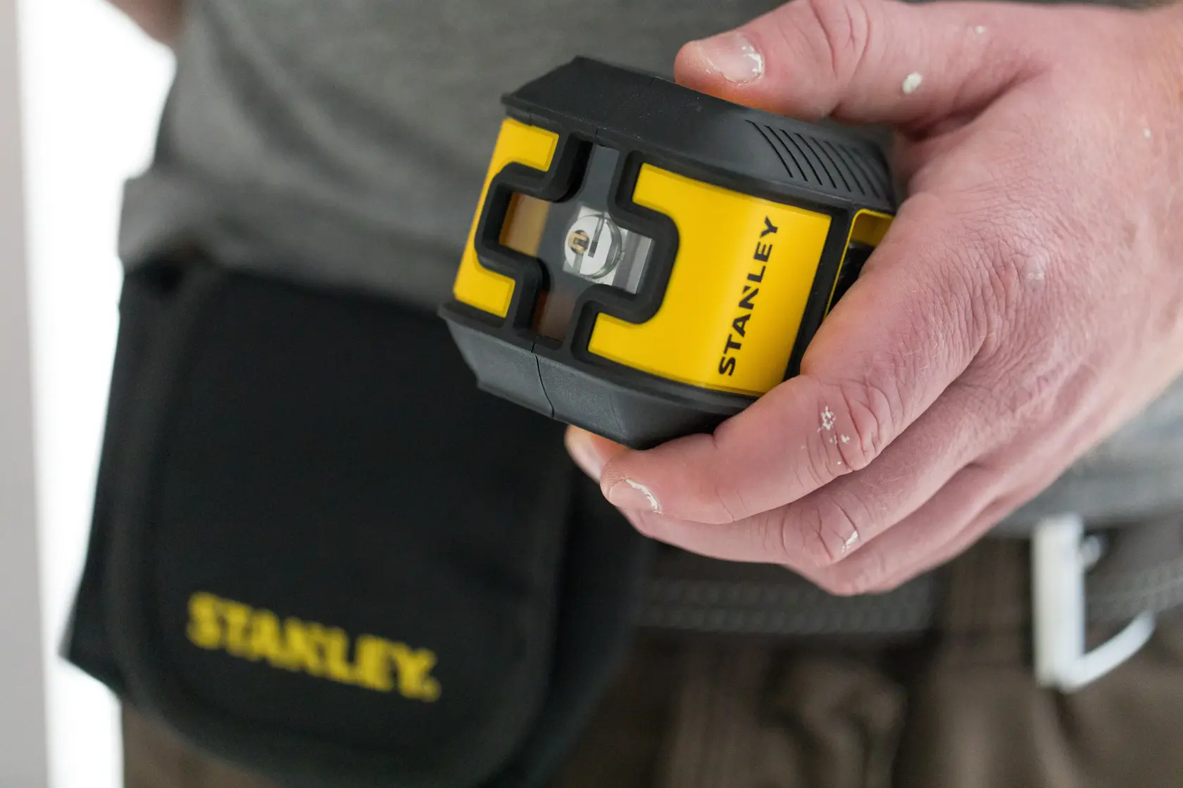 Fotografía del nivel láser amarillo y negro STANLEY STHT77498 sostenido en la mano, bolsa de herramientas visible al fondo.