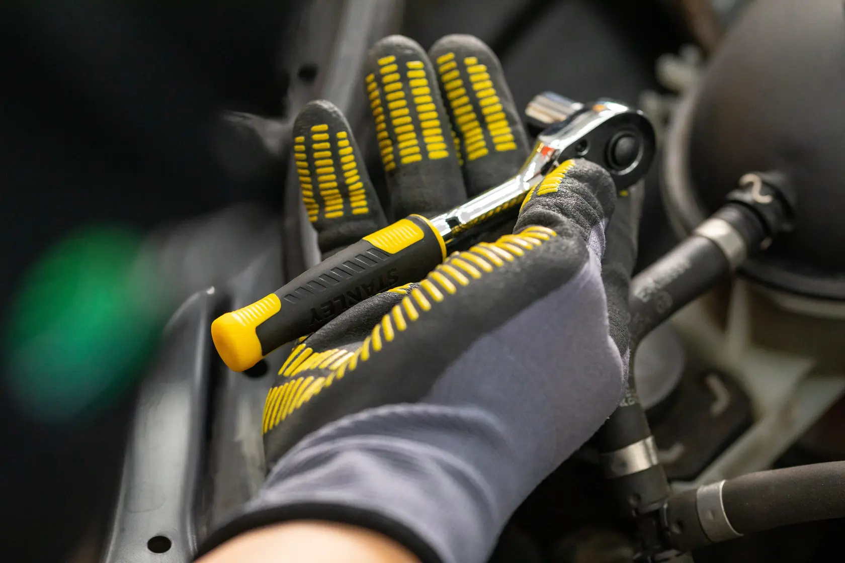 Llave de carraca STANLEY STMT82832-LA con mango negro y amarillo, sostenida por una mano enguantada en reparación automotriz.