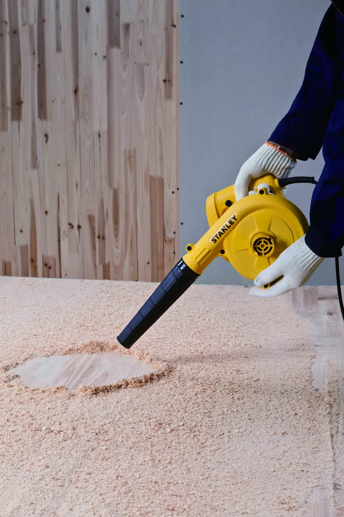 Fotografía del soplador eléctrico amarillo STANLEY STPT600-KR limpiando aserrín de una superficie de madera, ángulo frontal.