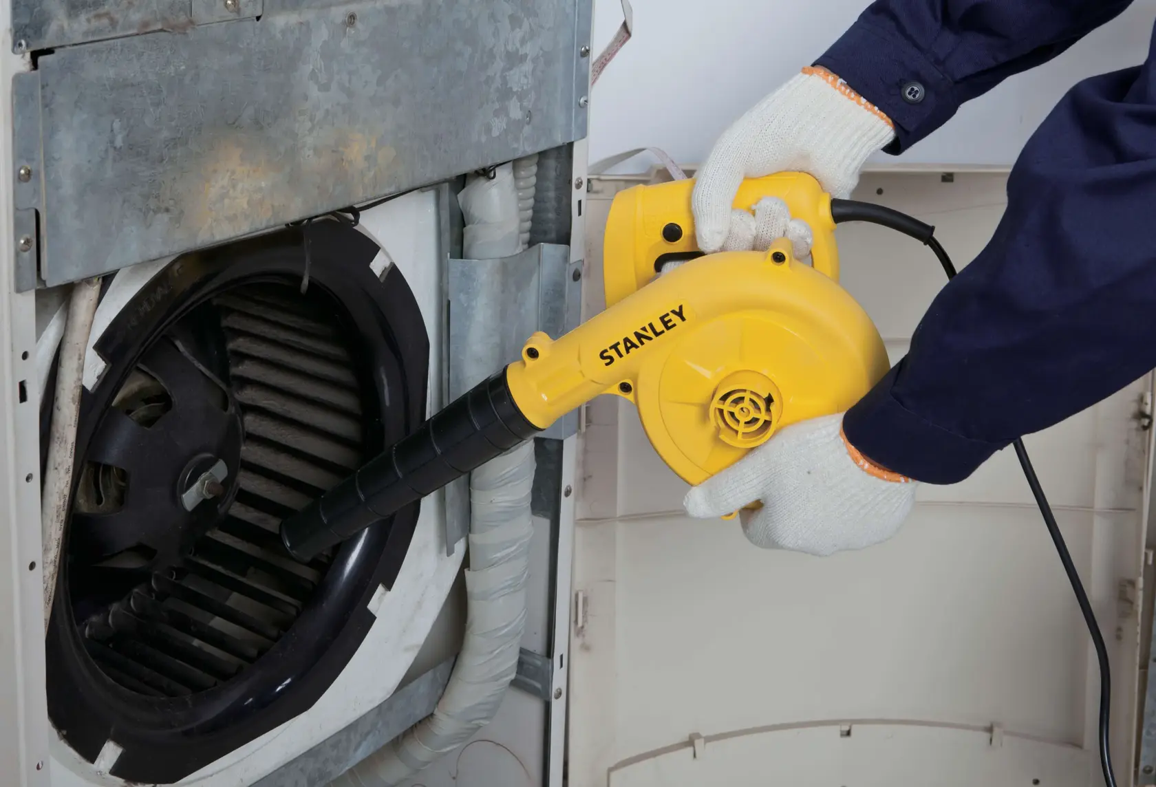 Fotografía del soplador eléctrico STANLEY STPT600-KR limpiando ventilas de máquina, cuerpo amarillo, boquilla negra.