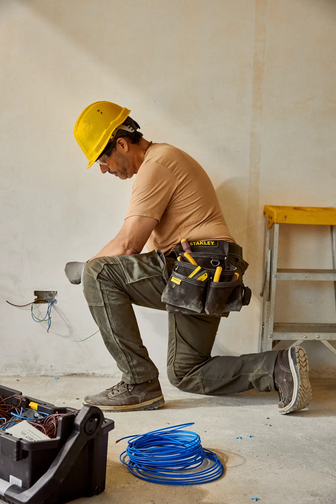 Fotografía de un trabajador con cinturón porta herramientas STANLEY STST1-80116, herramientas eléctricas visibles, arrodillado junto a una pared en construcción.