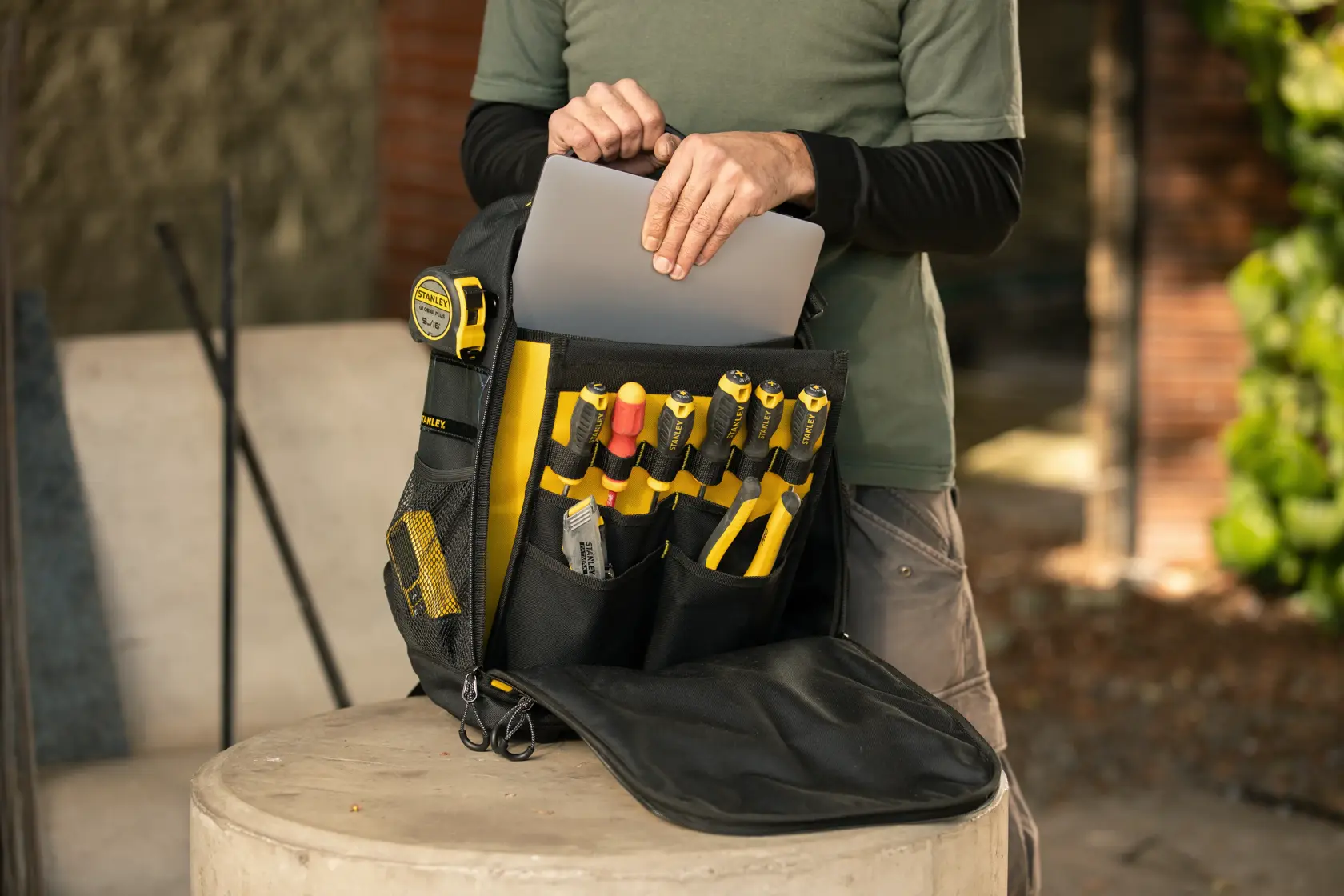Fotografía de mochila STANLEY STST515155LA con herramientas y cinta, persona guardando laptop en el bolsillo frontal.