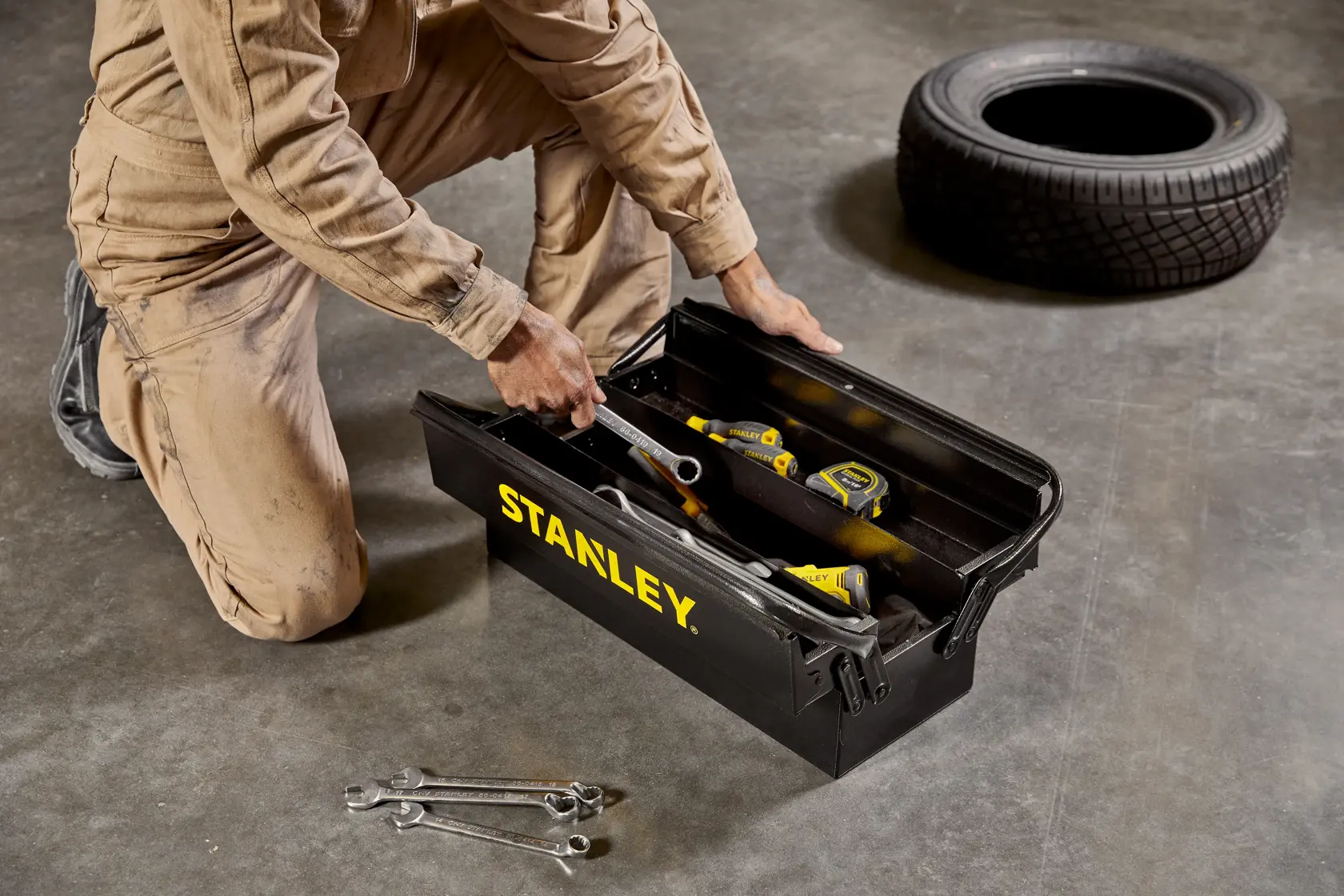 Caja de herramientas negra STANLEY STST98725LA con logotipo amarillo, abierta en el suelo con herramientas dentro, mecánico arrodillado cerca.